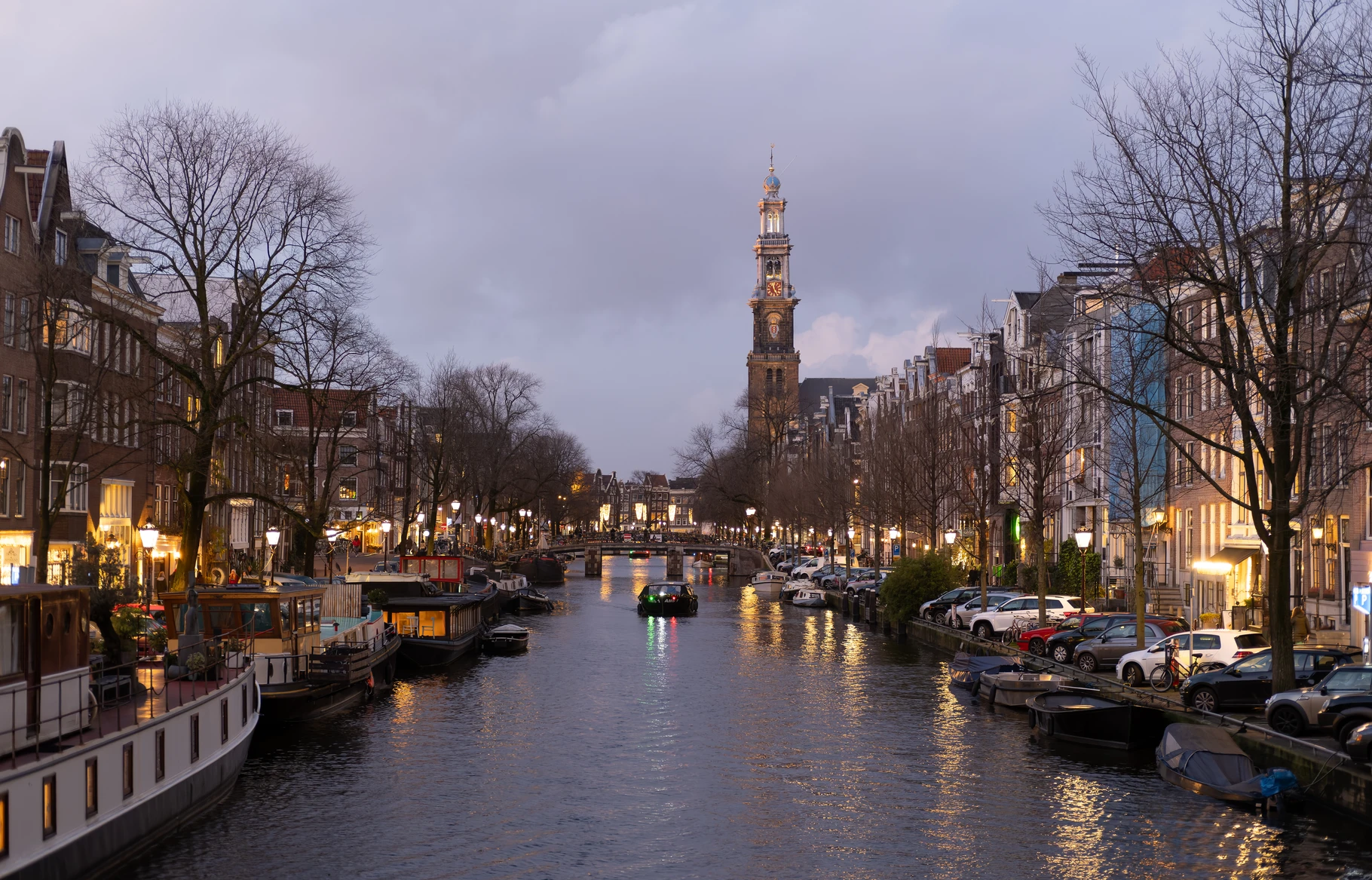 The Westerkerk tower rises above the Prinsengracht at dusk, seen down the canal from a Reestraat bridge