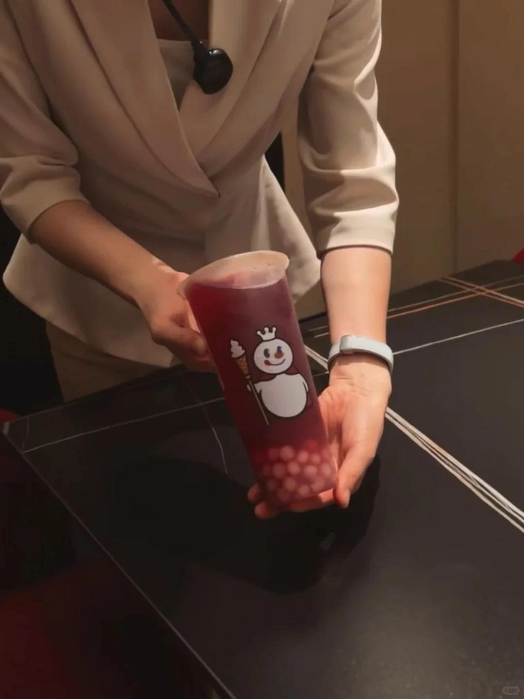 A server in a cream jacket presents a Mixue cup of Taro Ball Grape, Snow King mascot facing the camera, at a dark marble table