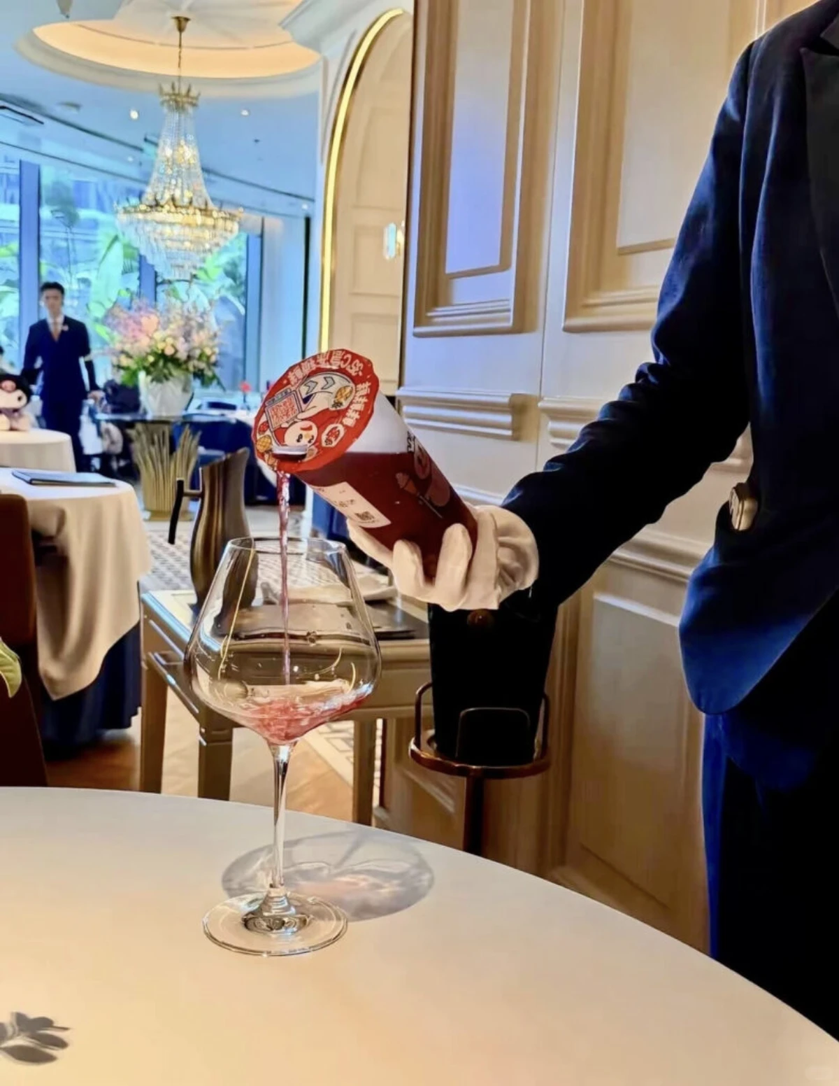 A white-gloved server in a navy jacket pours a purple Mixue drink from its branded cup into a large wine glass on a white tablecloth, in a hotel dining room with a chandelier