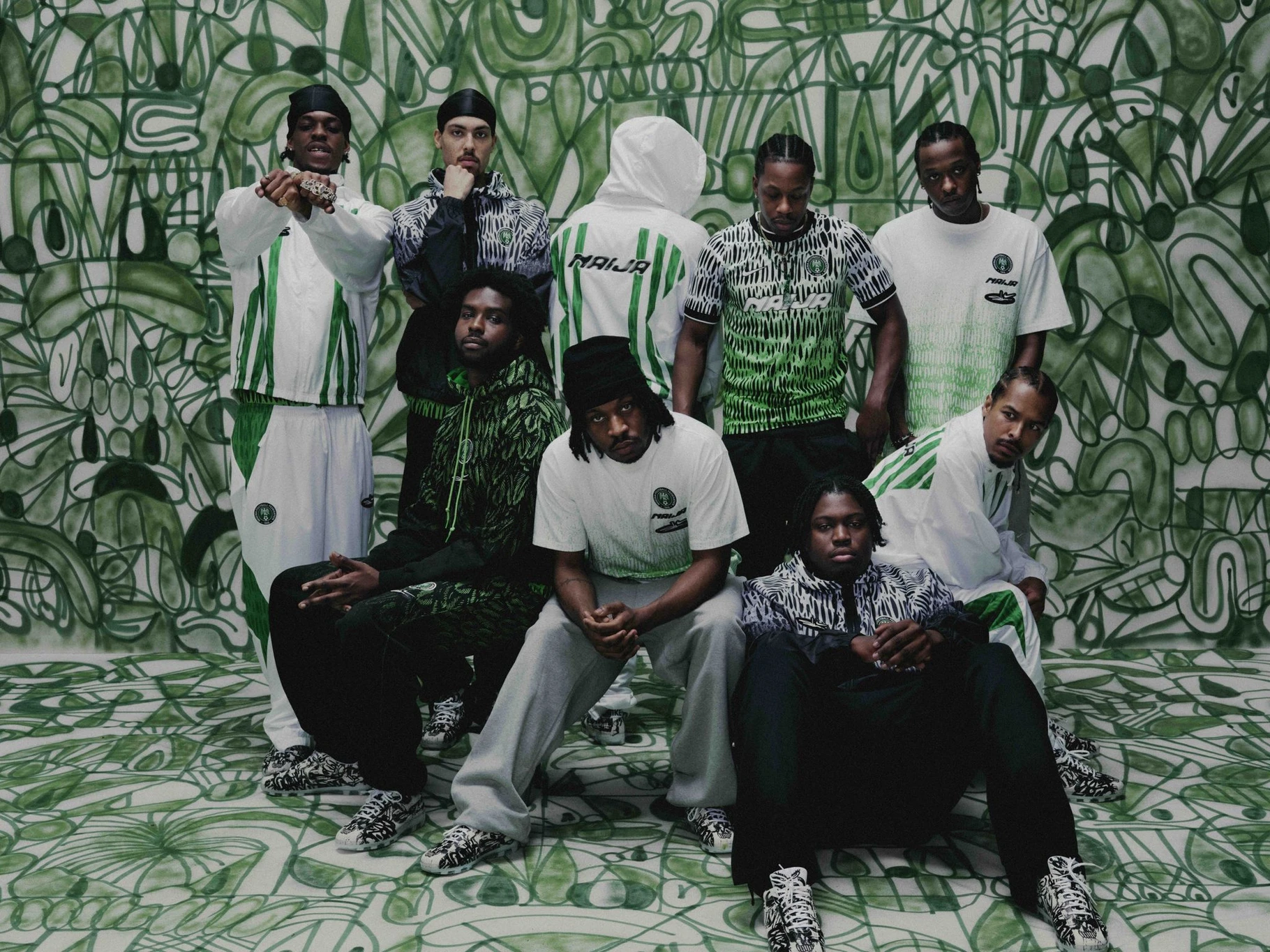 Nine models in Nike x Slawn Nigeria NAIJA jerseys and apparel pose inside a room painted with Slawn's green hand-drawn pattern