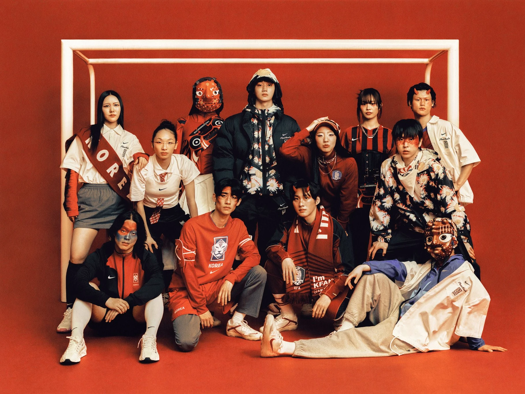 Twelve models in red and black PEACEMINUSONE x Nike Korea gear pose in front of a goal frame on a red set, two wearing traditional Korean masks